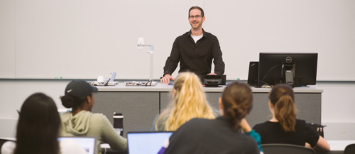 A professor speaks from behind a desk to students in a lecture hall.
