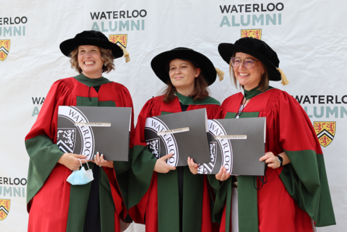 Three female PhD graduates stand dressed in traditional academic regalia while holding their diplomas.