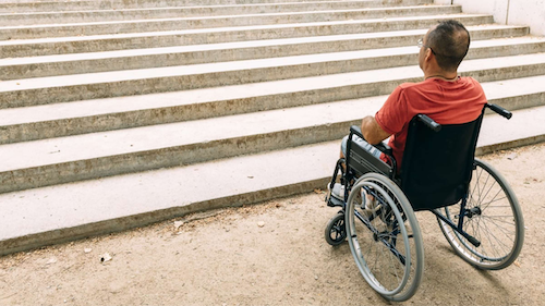 A man in a wheelchair contemplates an inaccessible set of concrete steps.
