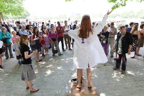 A scientist in a lab coat stands on a soapbox in a public park.