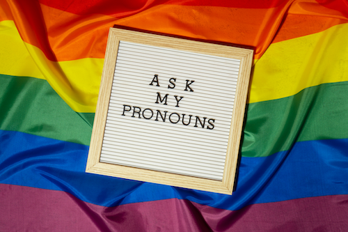 A framed sign saying &quot;ask my pronouns&quot; against a rainbow flag background.