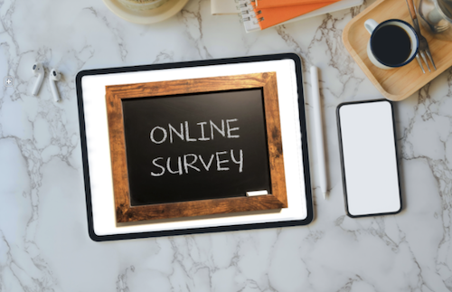 A tablet showing the phrase &quot;online survey&quot; sites on a table next to a phone and a cup of coffee.