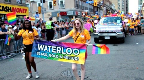 Two participants in the Pride Parade hold a sign that says Waterloo, Black, Gold and Proud.