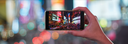 A person holds up a smartphone, which captures a vibrant city street on its screen.