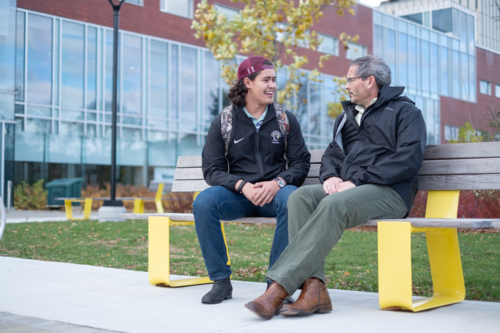 A student sits on a bench speaking with a professor.