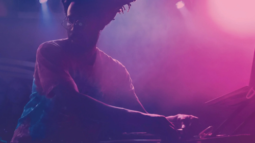 Chris Wilson performing behind the decks as he is lit by purple lights and fog.