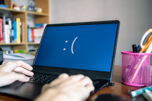 A person works on a laptop with the dreaded Blue Screen of Death and a sad emoticon on the screen.