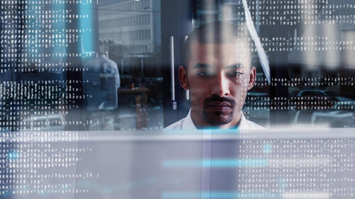 A Black man works at a computer as lines of code are superimposed over the image.