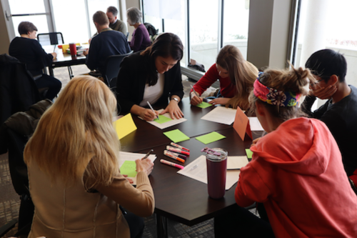 A group of people write on post-it notes while seated around a table.