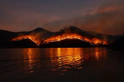 A forest fire burns on a mountainside as viewed from a lake.