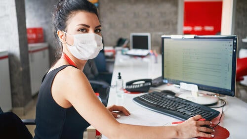 A woman sits at a desk wearing a mask.