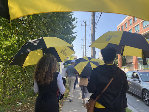 People holding University of Waterloo umbrellas march on a sidewalk.