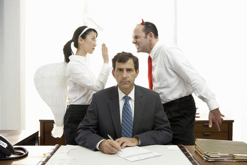 A man sits at a desk while an angel and a devil argue behind him.