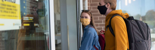 Two masked students enter a campus building.