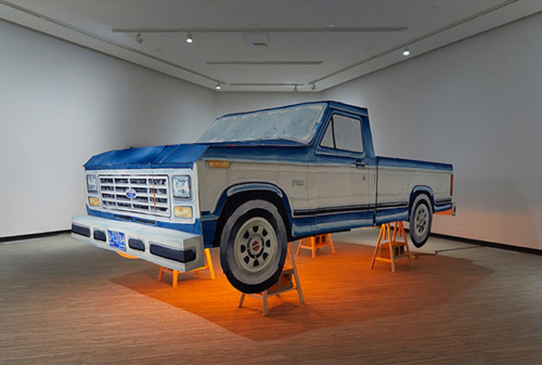 A life-sized papercraft pickup truck in white and blue stands upon four sawhorses, lit from underneath.