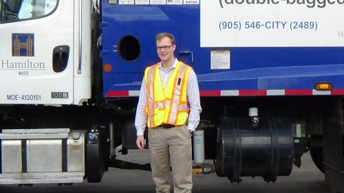 A student in a reflective vest stands in front of a Hamilton waste truck.
