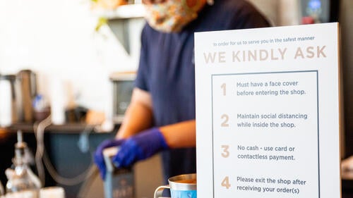 A coffee shop worker behind a sign asking patrons to wear masks and observe other rules.