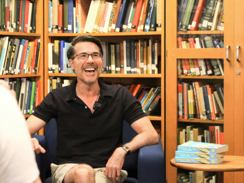 Brian Orend smiles as he sits in a chair with shelves covered in books behind him.