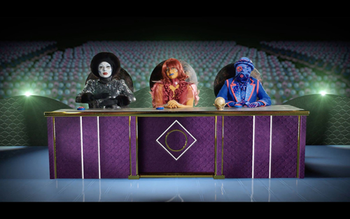 Three otherwordly characters acting as a judging panel sitting behind a desk.