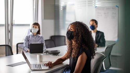 People in masks at a conference table watch a presentation.