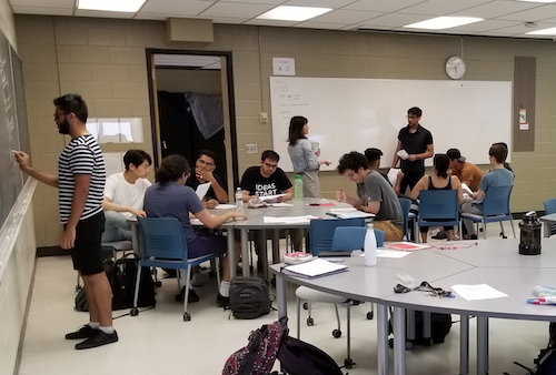 Students in an active learning environment with tables arranged in pods and whiteboards.