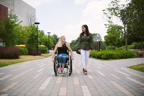One woman in a wheelchair and another walking alongside her on campus