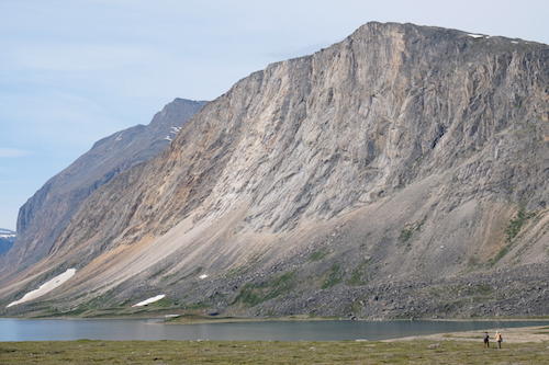 Two people are dwarfed by an imposing cliff face on the shore of a lake.