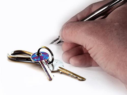 A person signs a document with a pen with house keys in the foreground.
