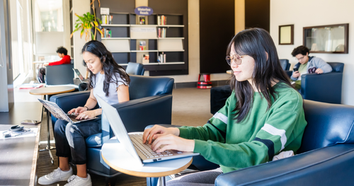 Two women work on laptops in a library setting.