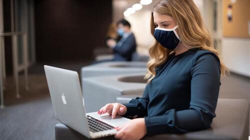 A person in a mask works on a laptop.