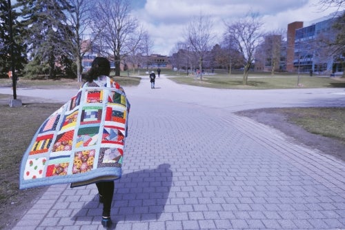 A student wrapped in a traditional quilt walks along a campus pathway.