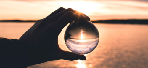 A person holds a glass sphere up to a lake, showing an inverted reflection.