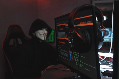 A hooded hacker in a Guy Fawkes mask looms over a computer screen.