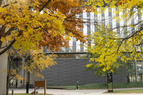 A person walks in front of the facade of the Quantum-Nano Centre with autumn leaves overhead.