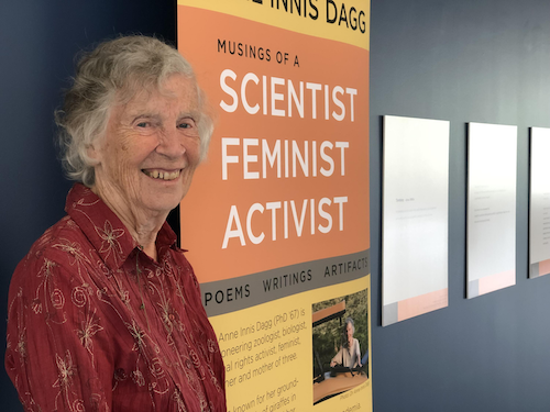 Dr. Anne Dagg standing next to a banner that reads &quot;Musings of a Scientist, Feminist, Activist.&quot;
