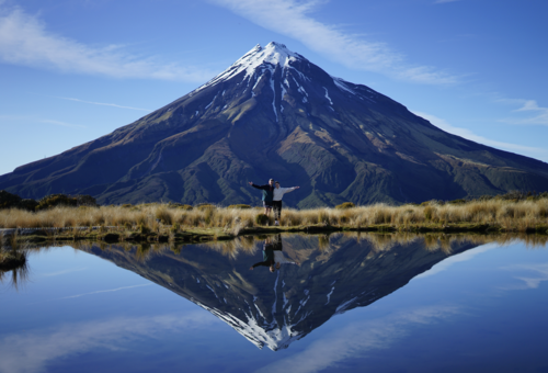 Two people pose in front of Mount Taranaki in New Zealand reflected mirror-style in a lake.