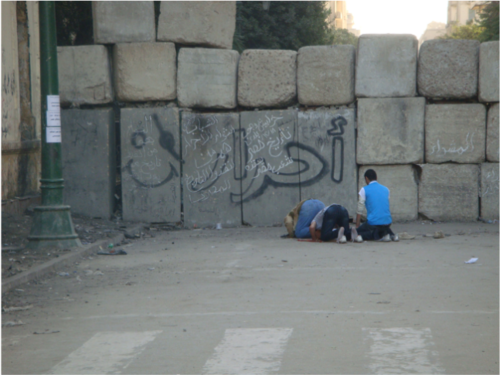 Men kneel in prayer before a brick wall covered in Arabic script.