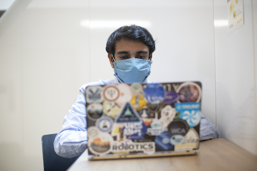 A man wearing a mask works on a laptop covered in stickers.