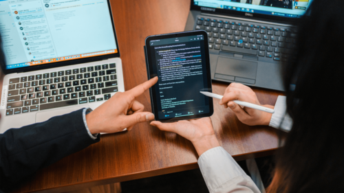 Two people consult a tablet with their laptops open on a table in front of them.