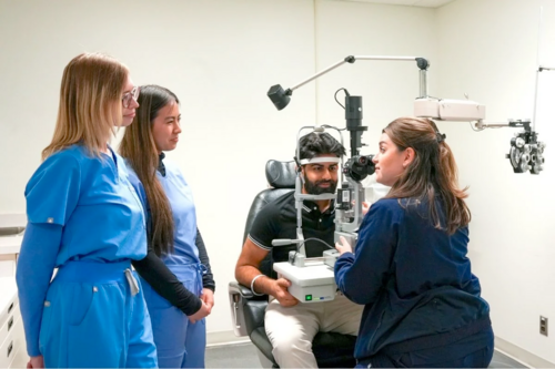 An optometry instructors demonstrates how to give an eye exam to a patient in a chair while two students look on.