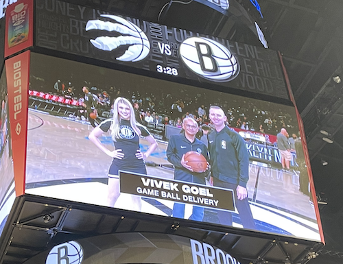 A shot of the court's video screen showing President Vivek Goel delivering the game ball to an official while a cheerleader looks on smiling.
