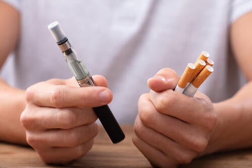 A person holds a vape pen in one hand and a bundle of cigarettes in the other.