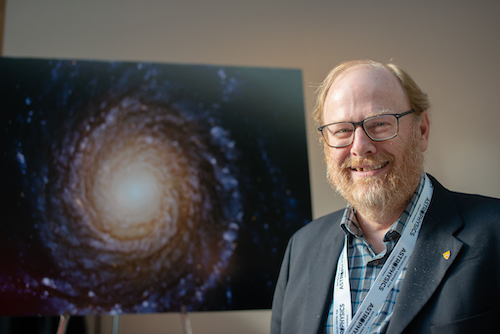 Dr. Mike Fich stands with a painting of a galaxy or other stellar object.
