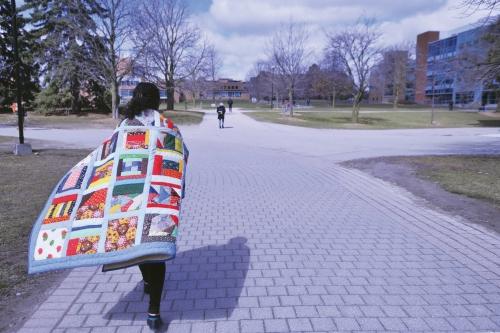 A woman walks on campus with a quilt over her shoulders like a cape.
