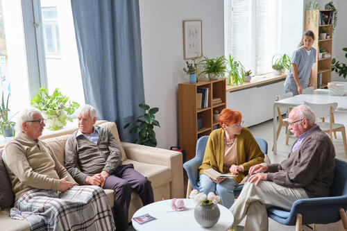 Residents at a long-term care home sit on couches while a personal support worker works in the background.