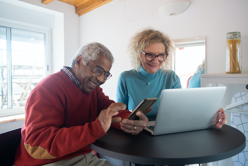 An elderly man and woman smile as they look at their phone and laptop.
