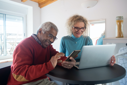 Two elderly people smile and laugh as they interact with their smartphone and laptop.