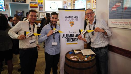 Three Waterloo alumni hold up pennants and smile next to an Alumni and Friends zap banner.