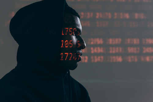 A stereotypical computer hacker wearing a black hoodie as red digital numbers are projected on his face.