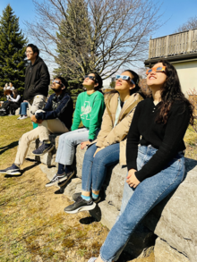 Students wearing eclipse glasses look up at the solar eclipse with the Grad House in the background.